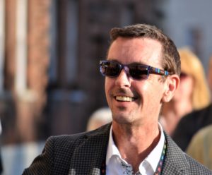 Picture shows Andy Hunt, in a jacket and white shirt, smiling and wearing sunglasses. He is outside and the sun is on his face. 