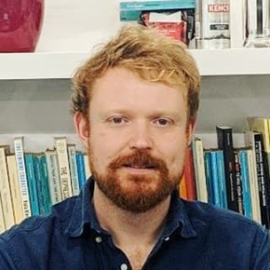 Image of Fred Cooper; he is standing in front of a bookcase with books, wears a blue shirt and has reddish hair and a beard. He is looking directly into the camera.