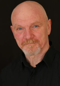 Picture shows a colour portrait of Keiron, who is positioned slightly side on to the camera. He is wearing a black shirt. Neutral expression.