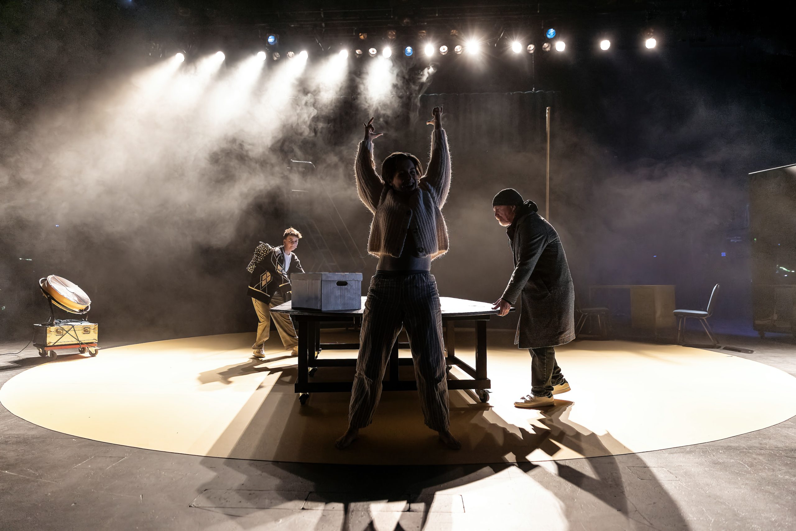 A photo of the production. A figure stands with their arms raise dramatically in the air in the foreground. White lights and smoke jet from the top of the image. In the background, two figures move a table and a box, in preparation for the next scene.