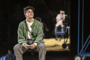Luca sits in a green jacket and grey trousers, holding their sketch pad
