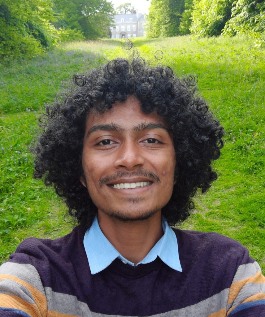 Abhishek has brown skin and black curly hair. They are taking a selfie in front of a beautiful springtime wood and field, everything behind them is green. They are smiling and are wearing a stripy jumper.