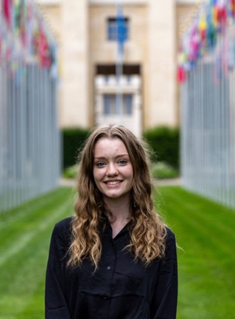 Abby is outside the UN Palais Des Nations, she has long wavy dark blonde hair and is wearing a black blouse. She is smiling. The flags of the UN Nations are displayed on poles behind her.