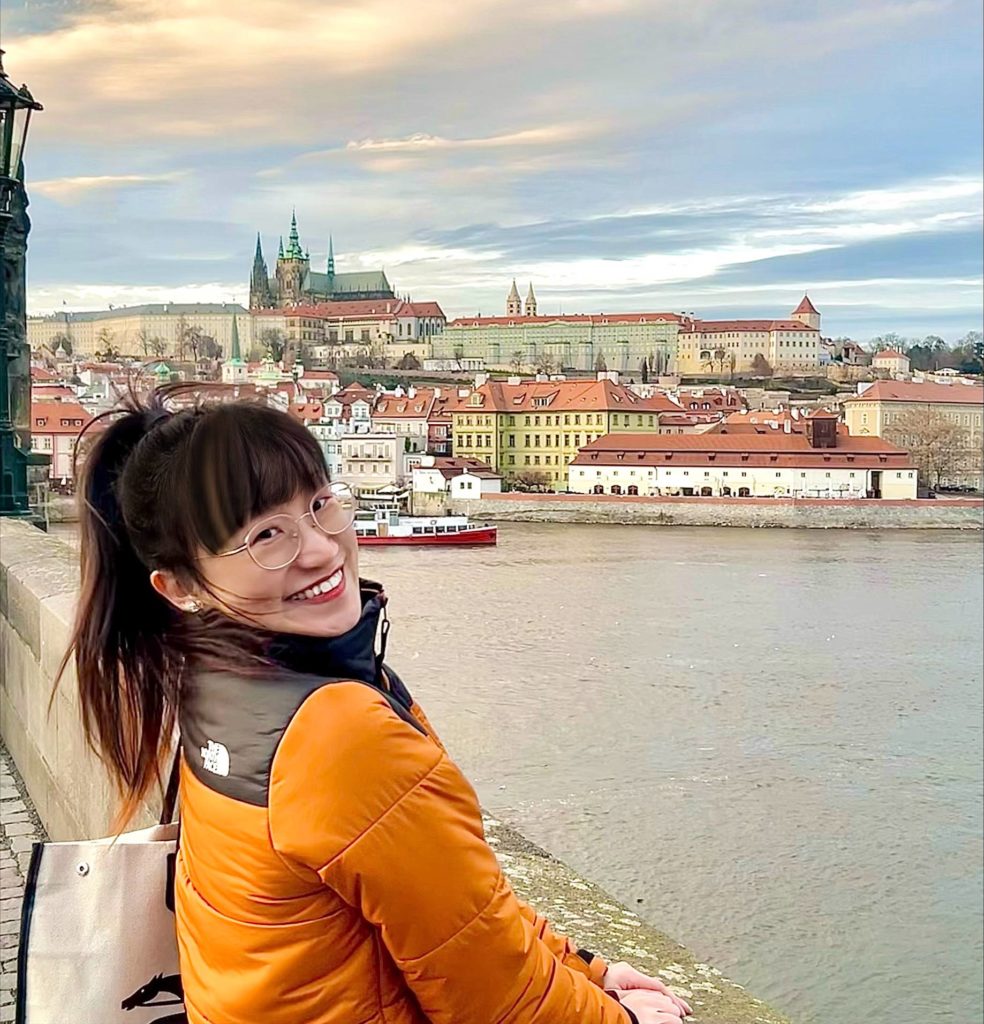 Natalie is standing on a bridge in Prague. Natalie has long dark brown hair, she is wearing glasses and smiling. She is wearing an orange jacket. There are boats on the river below her.