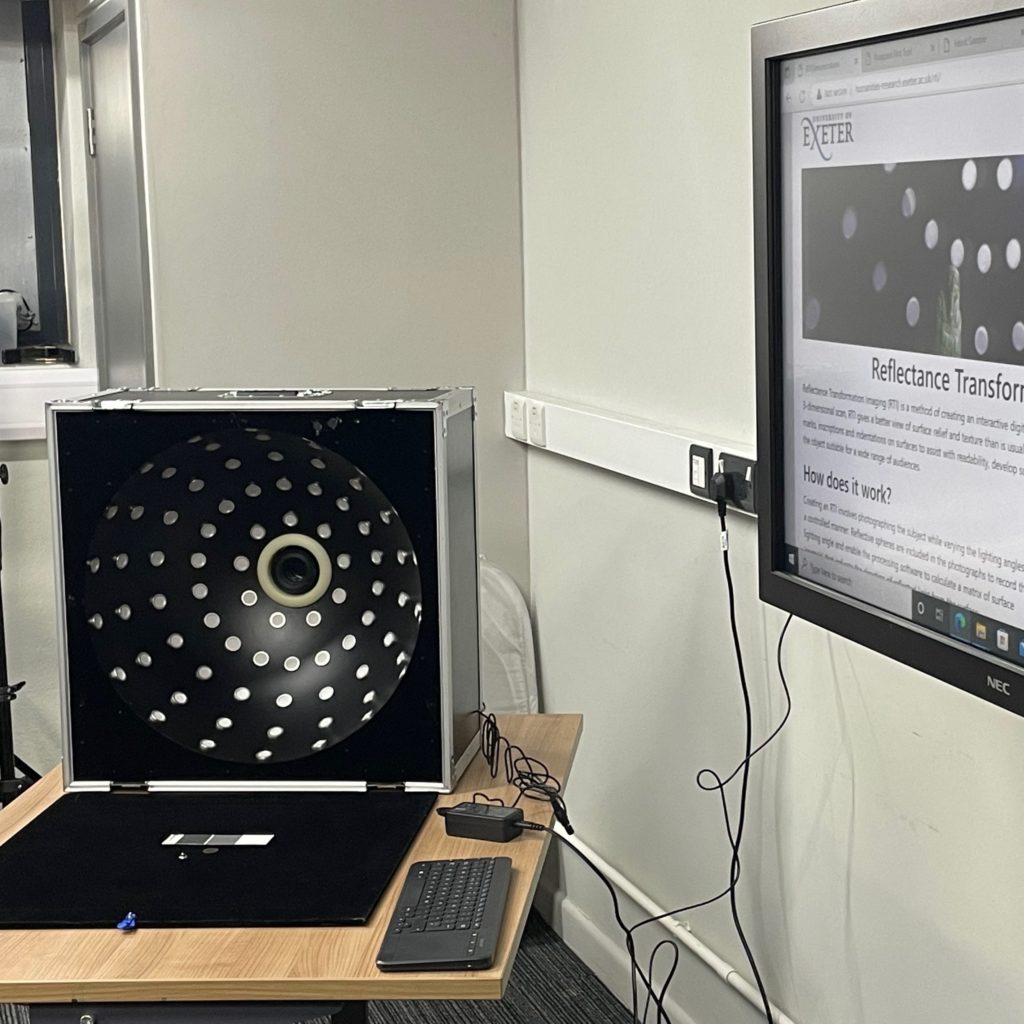 An open Reflectance Transformation Imaging machine on a desk next to a screen displaying a University of Exeter website 