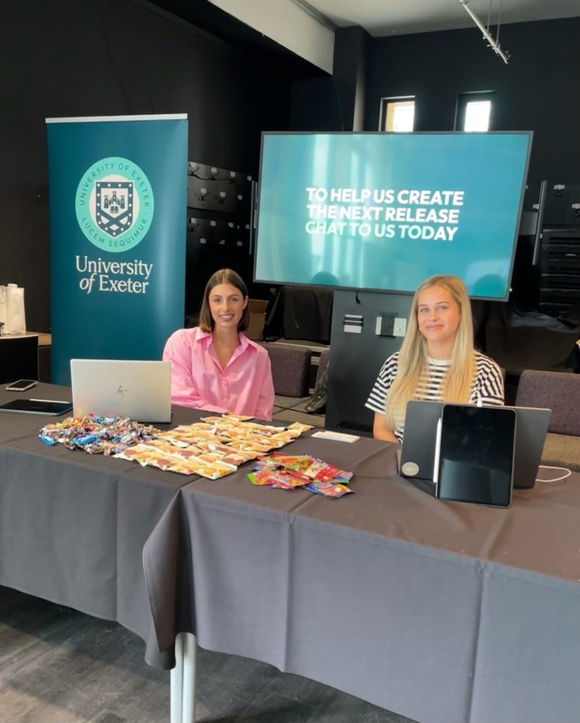 Two woman sat at a table in front of a University of Exeter banner and TV with promotional video playing