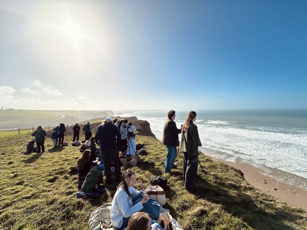 A group of people sat and stood on a coastal cliff, looking out to sea