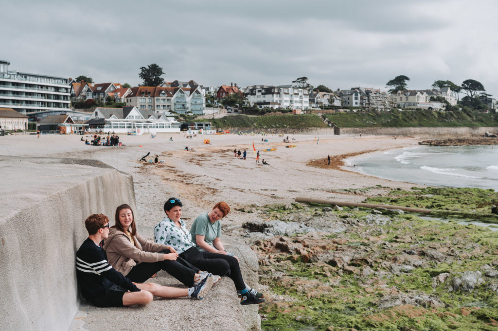 A group of four students sat against the sea wall in the sunshine. Behind them is gylly beach in Falmouth with views towards the rockpools, cafe and the sea