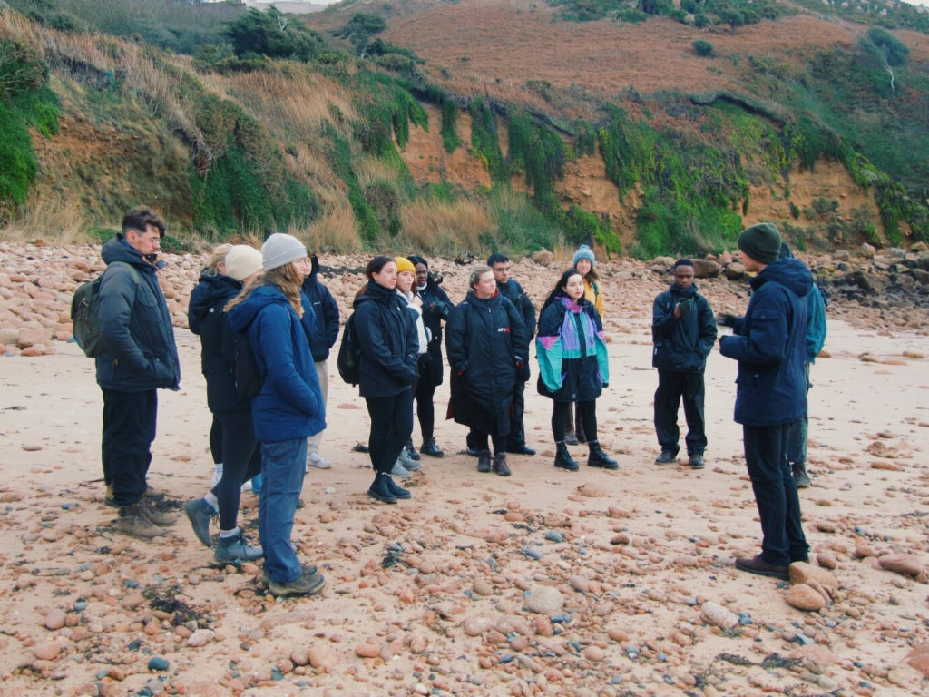 MSc students on the field course to Jersey, stood on a beach in front of some cliffs listening to a discussion.