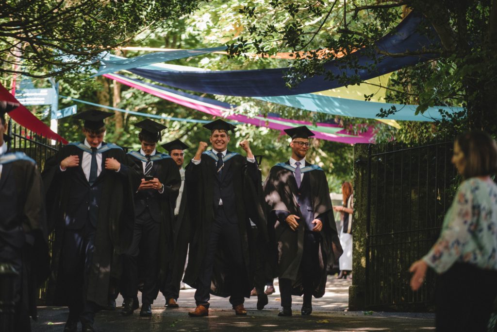 Graduates leaving their graduation ceremony In their gowns. They are walking down a lane in the sunshine beneath colourful banners hanging between trees. A graduate in the centre is celebrating with his fists in the air and a big smile.