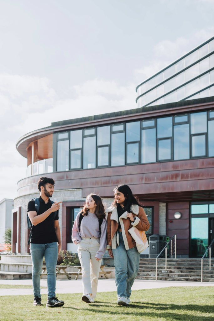 University of Exeter Penryn Campus in Cornwall. Three students walk across grass in the sunshine, in front of the Stannary and Sustainability Cafe.