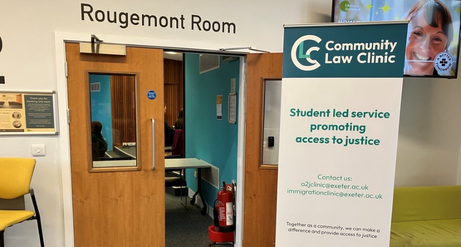 A tall pull up display sign reads Community Law Clinic. Student led service promoting access to justice. Contact us: a2jclinic@exeter.ac.uk, immigrationclinic@exeter.ac.uk. Behind is the entrance to a room with one door open. Above the door are the words Rougemont Room.