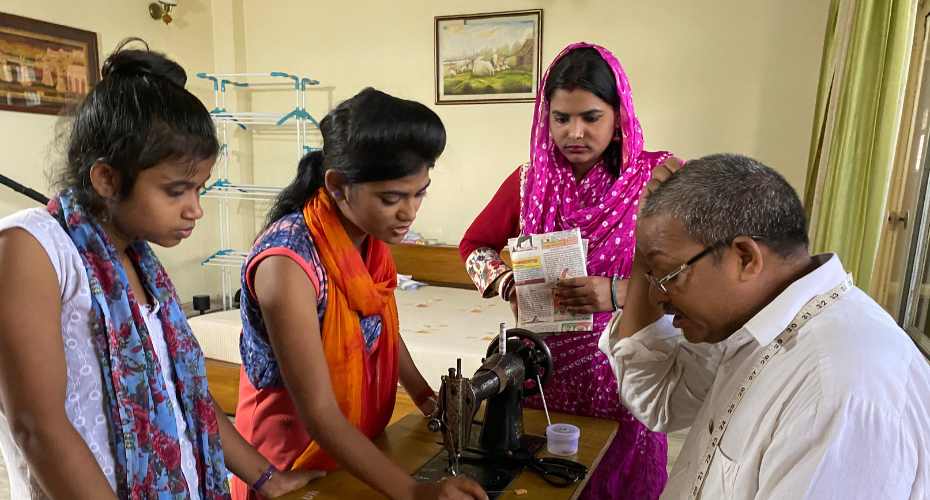 Three people in a tailor's workshop in India, watching a tailor at a sewing machine. One woman is wearing pink, another orange and the third is dressed in blue. The tailor is wearing a white shirt.