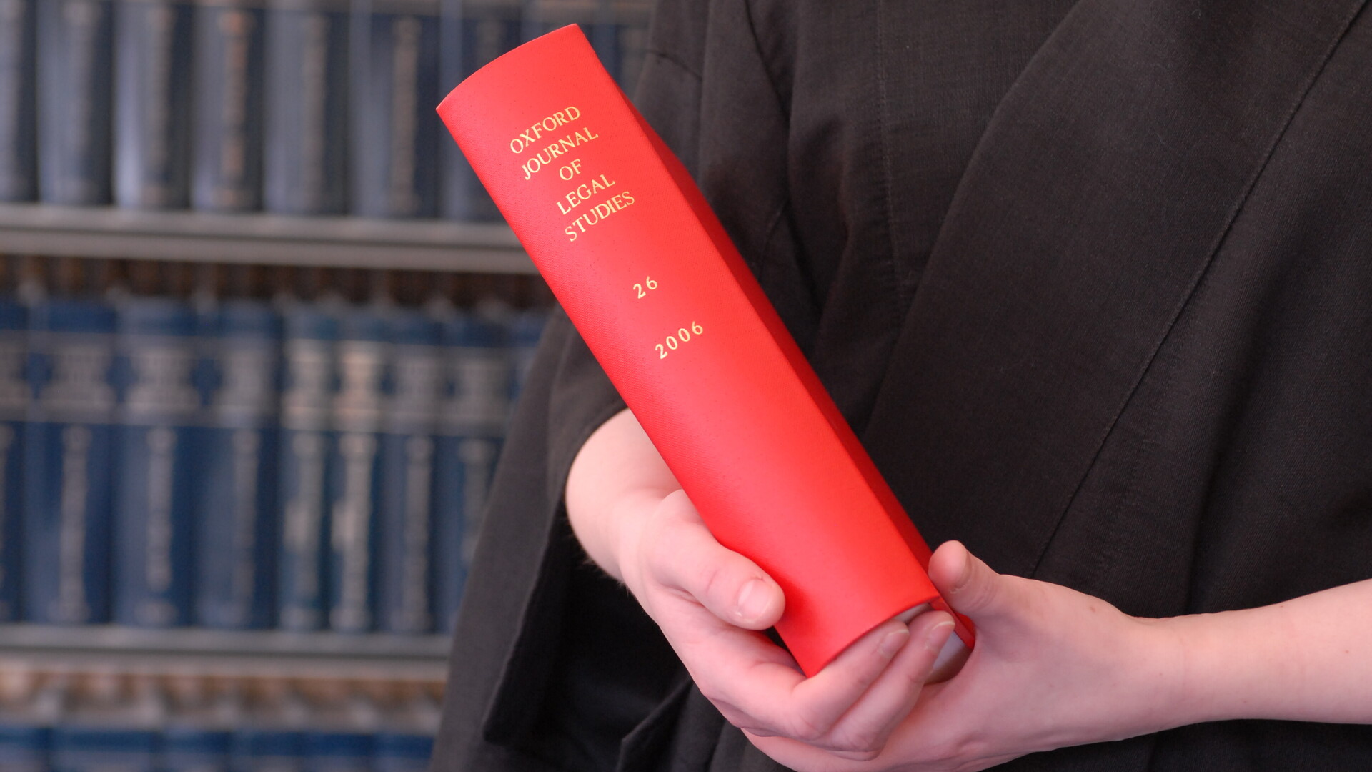 a red book held by two hands. A bookcase is in the background