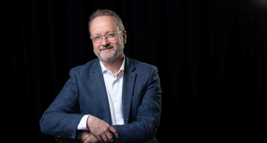 Head and shoulders photo of Professor Moorhead. He is wearing a blue jacket and white shirt and has his hands in front of him, slightly folded. the background is black.