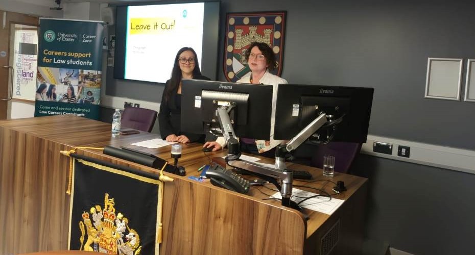 Two people stood at the front of the Moot Court for the Canadian Law Careers Development Event. They are in front of a large brown desk with two screens in front of them.