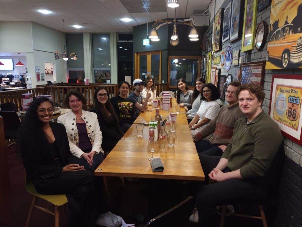 A group from the event sat at long table in one of the campus eating venues. The people are all turned towards the camera and smiling. On the wall are various decorative pictures.