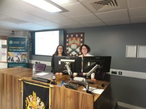 Two people stood at the front of the Moot Court for the Canadian Law Careers Development Event. They are in front of a large brown desk with two screens in front of them.