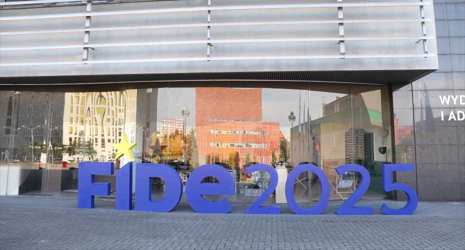 Large letters displayed outside of a grey building which has horizontal metal features and reflective glass in which you can see other buildings. The letters spell out FIDE 2025.