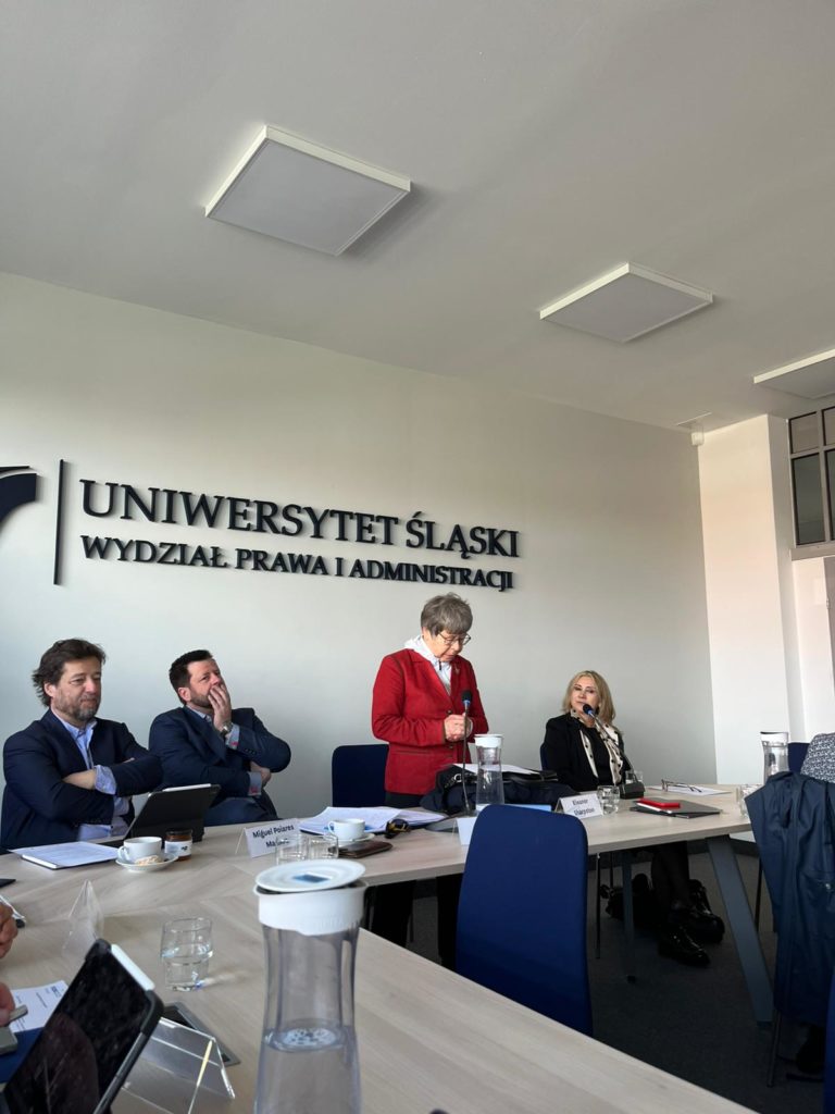 People gathered in a room for a conference with laptops, water bottles and the words Uniwersytet Śląski on the wall.