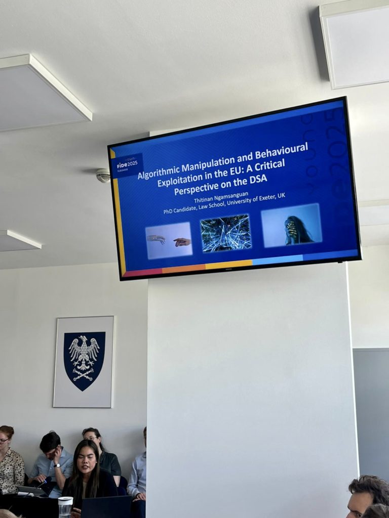A television screen displaying a slide saying 'Algorithmic Manipulation and Behavioural Exploitation in the EU: A Critical Perspective on the DSA’, Thitinan Ngamsanguan, PhD Candidate , Law school, University of Exeter. You can also see some of the attendees at the presentation.