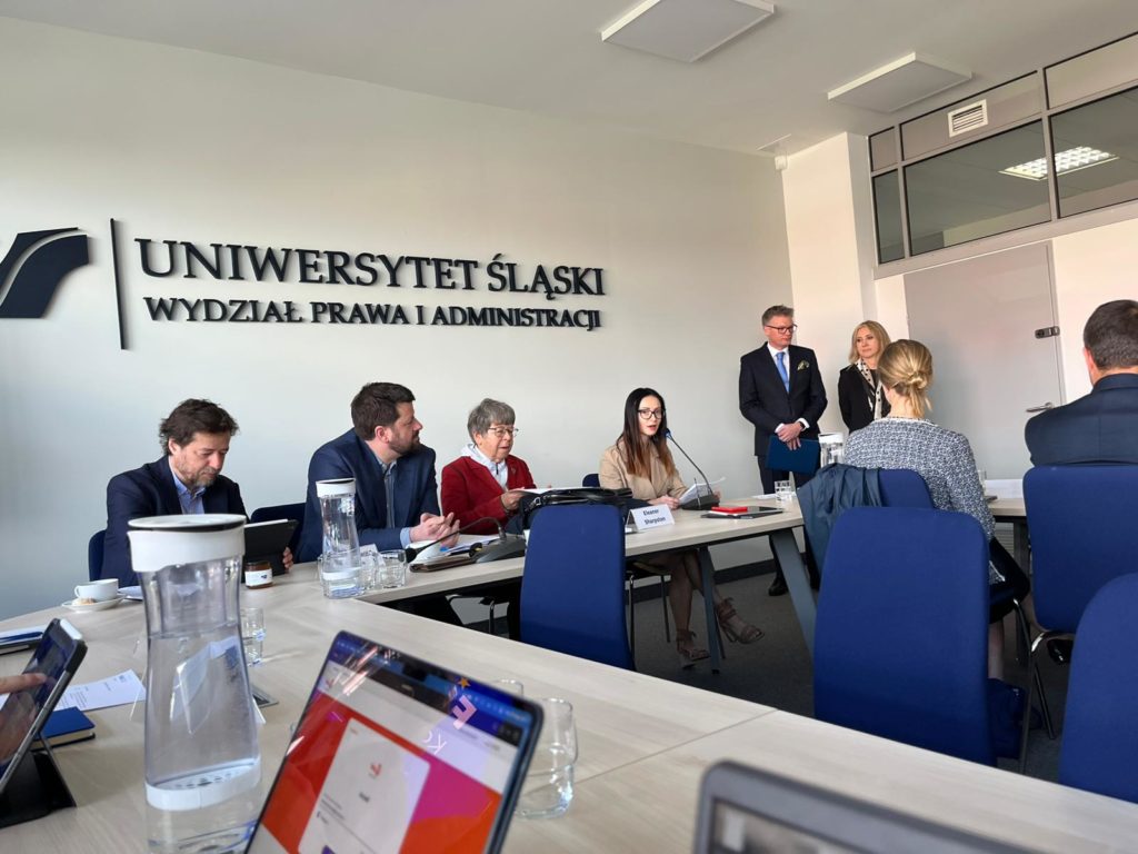 People gathered in a room for a conference with laptops, water bottles and the words Uniwersytet Śląski on the wall. Two people are standing and three are seated listening to them speak.