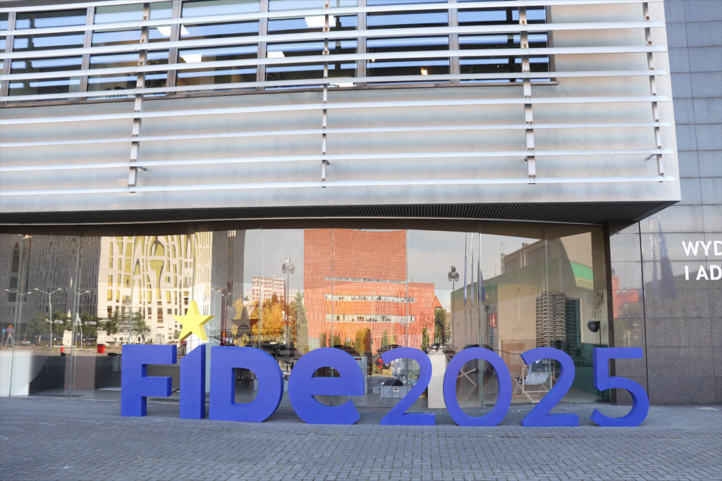 Large letters displayed outside of a grey building which has horizontal metal features and reflective glass in which you can see other buildings. The letters spell out FIDE 2025.