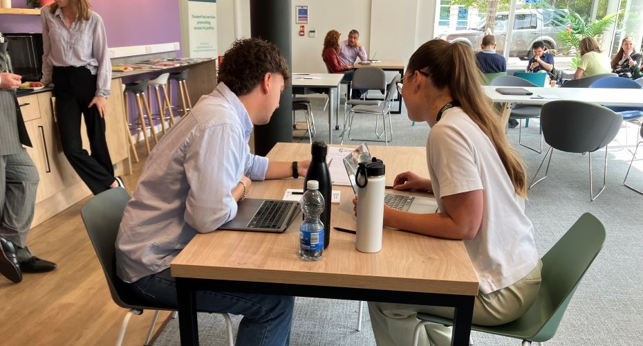 Two students sat at a desk discussing work with laptops, pen, papers, water bottles. 