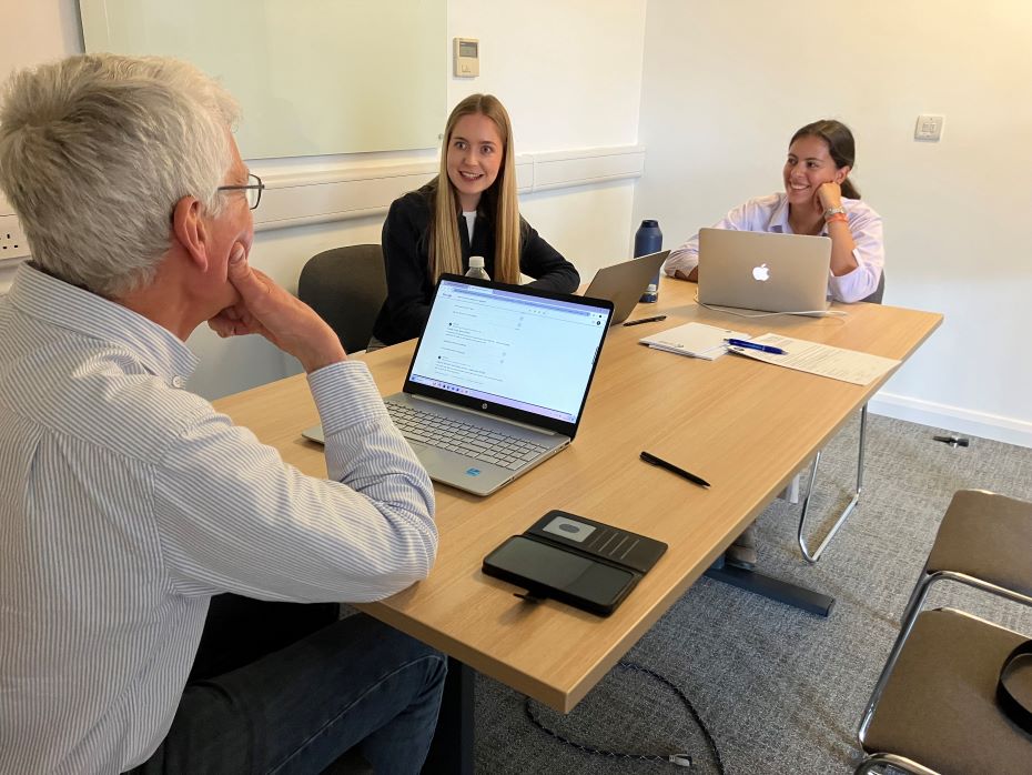 Two students and a supervisor sat at a desk discussing work with laptops, pen, papers, water bottles. 