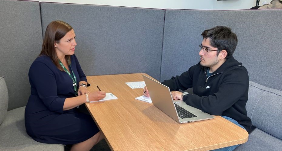 One student and a supervisor sat at a desk discussing work with laptops, pen, papers. 