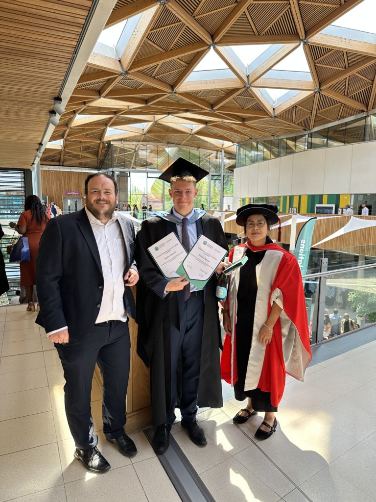 Prize winner (Baker Prize for Company Law and Malcolm Mead Employment Law Prize) Matthew Barrett stood between Richard Bowyer and Amy Man. They are wearing graduation robes and Matthew is holding a certificate. The backdrop is a curved wooden ceiling with triangular inset windows.
