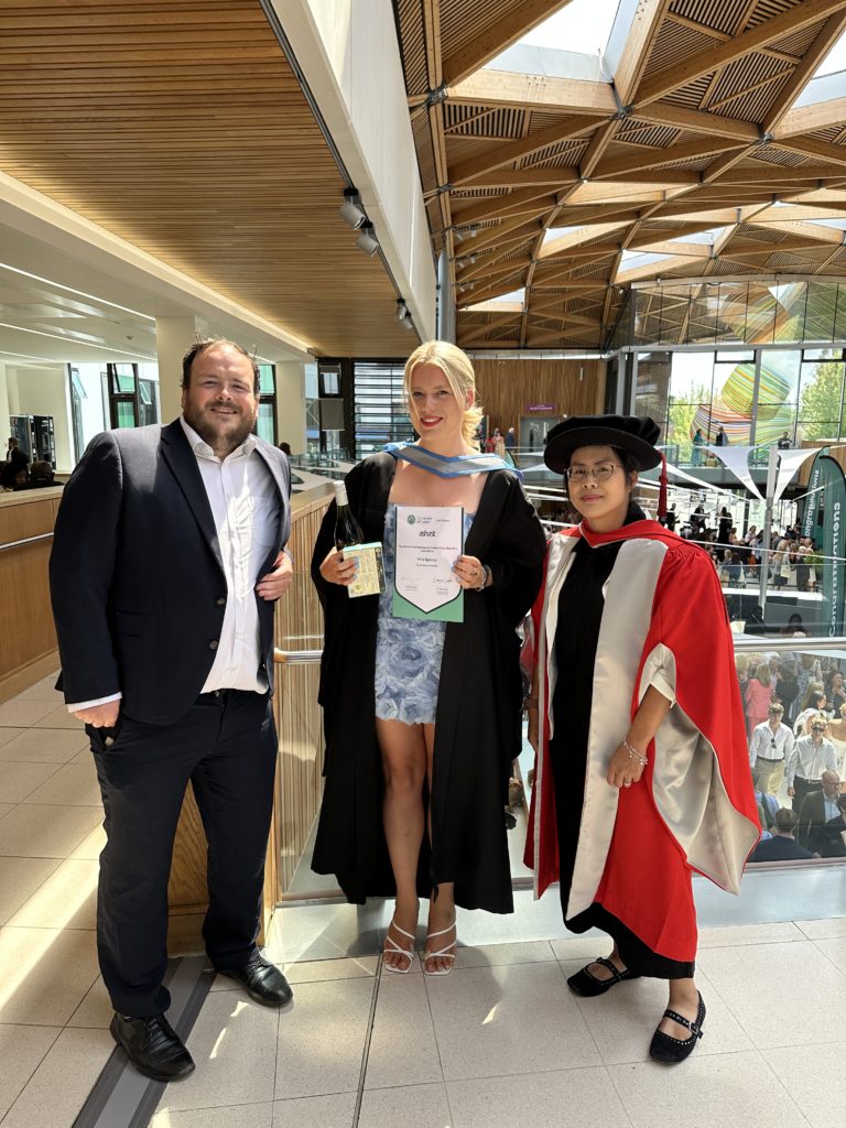Prize winner (The Ashurst Outstanding Law Student Prize) Mila Spence stood between Richard Bowyer and Amy Man. They are wearing graduation robes and Mila is holding a certificate. The backdrop is a curved wooden ceiling with triangular inset windows.