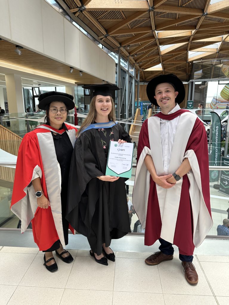 Prize winner (IPAN Prize for intellectual property) Caitlin Mainwaring stood between Amy Man and David Yuratich. They are wearing graduation robes and Caitlin is holding a certificate. The backdrop is a curved wooden ceiling with triangular inset windows.