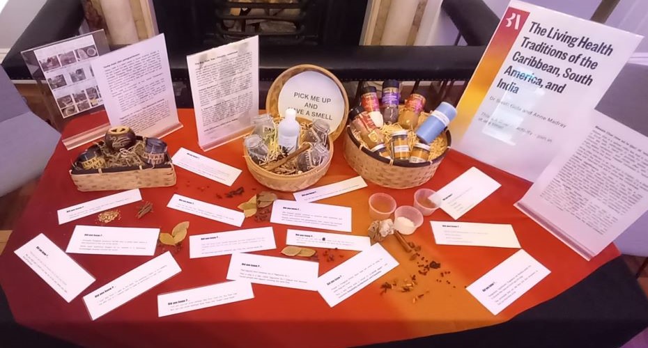 A table displaying a few baskets of items such as spices and herbs in jars. There are pieces of paper on the table next to small scatterings of leaves and spices. There is a display sign on the table which reads 'The Living Health Traditions of the Caribbean south America and India'. Another sign reads, Pick me up and have a smell.