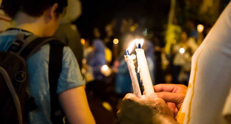 Two white lit candles being held by two hands. In the blurred background there are people also holding candles.