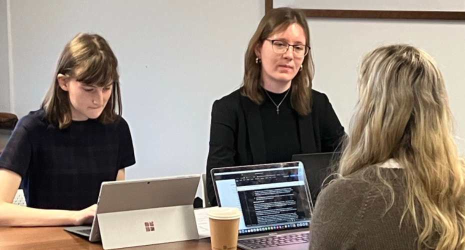 three students sat at a table with laptops in front of them. Two are engaged in conversation and one is typing on the laptop.