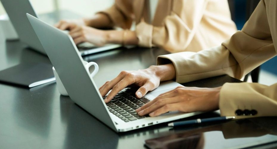 a photo of a laptop. There are two hands typing and in the background there is another person also with their hands on a keyboard.