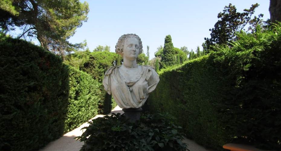 An ornate bust statue sits above a lush green hedge and is flanked by hedges on either side. There are tall trees in the background.