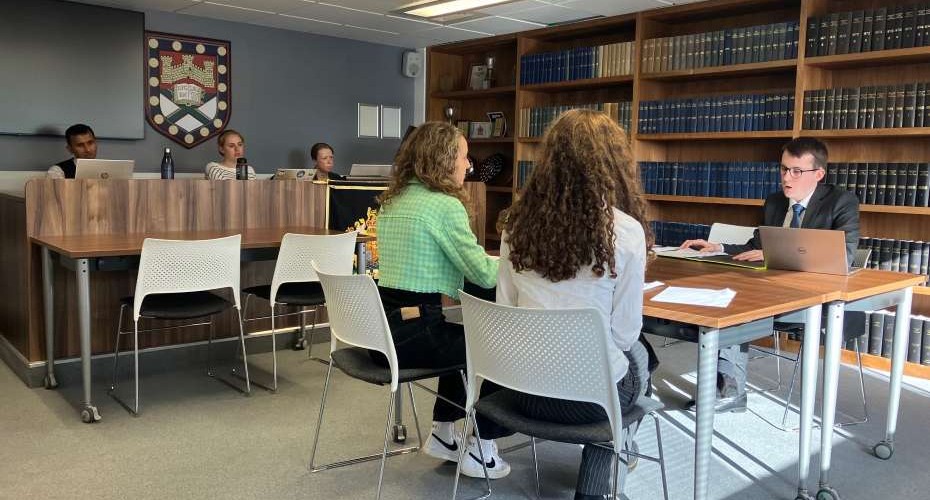 A fictitious courtroom setting. Two students are negotiating with a person on the other side of the table. There are three people sat observing behind a large wooden desk. There are many books on the shelves behind.