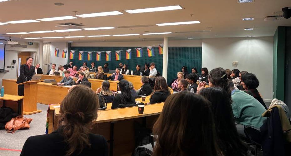 A room set up for Mooting with students sat at desks in rows. One person is stood up addressing the room. There is a row of penant rainbow flags strung along one wall.