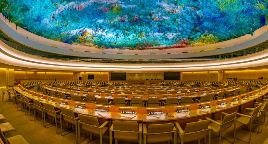 The interior of the United Nation's Palais des Nations in Geneva. It has a ornate multi coloured circular ceiling and the floor has circular rows of chairs, curved table and microphones.
