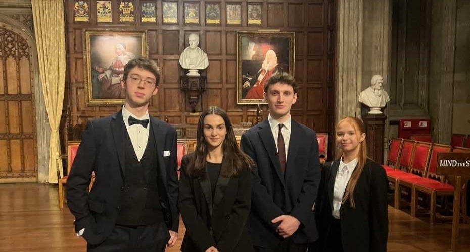 Four people dressed in formal clothing standing together in an ornate hall with wooden panelled walls, red upholstered chairs, and statues on plinths. Two large framed portraits hang on the back wall, and the room features carved doors, tall curtains, and polished wooden floors