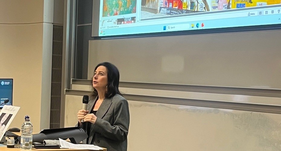 Human Rights lawyer is stood in front of a large display screen and next to the lecture presentation desk.