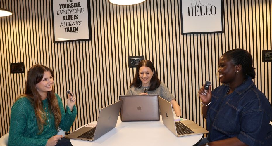 Three students are sat around a table with their laptops in front of them. They are holding small microphones as they chat together.