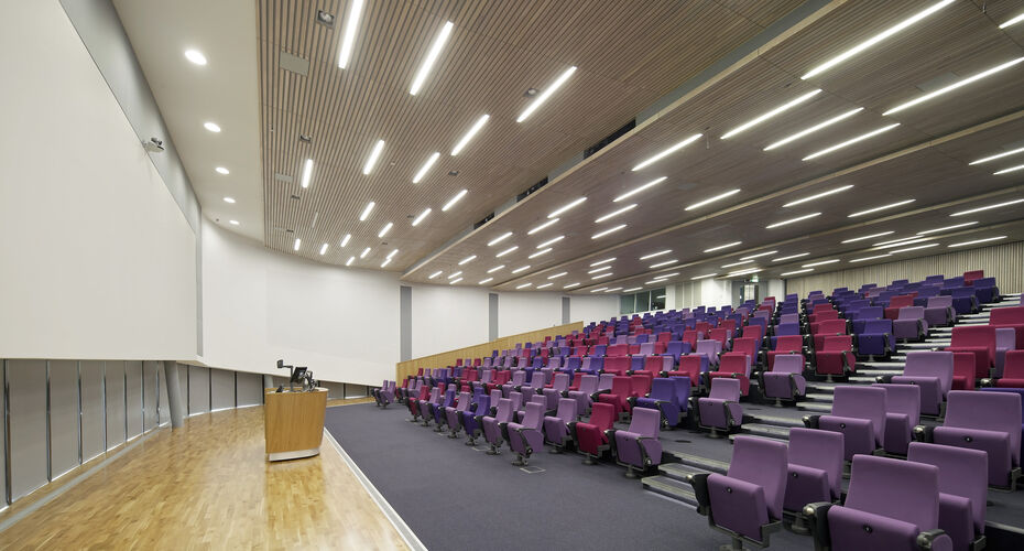 A lecture theatre with rows of purple and pink seats, strip lighting overhead and a podium at the front