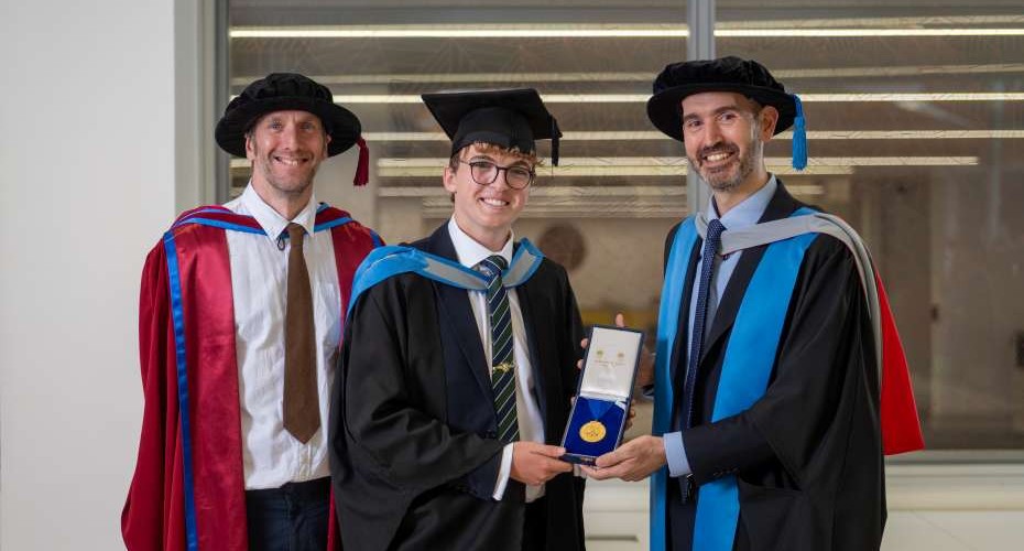Three people in academic dress pose indoors during a graduation ceremony. The person in the centre wears a graduation cap and gown and holds an open presentation case containing a medal, which is being presented by the person on the right. The person on the left stands beside them in a red academic robe. A windowed interior forms the background.