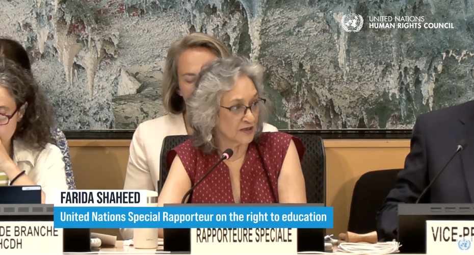 A woman sat at a desk in the UN speaking into a microphone, with other attendees in the background