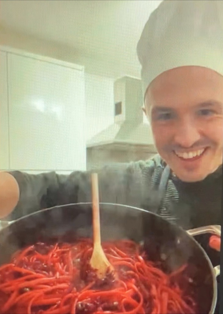 A smiling David yuratich wears a chef hat and holds a pan of pasta up to the camera