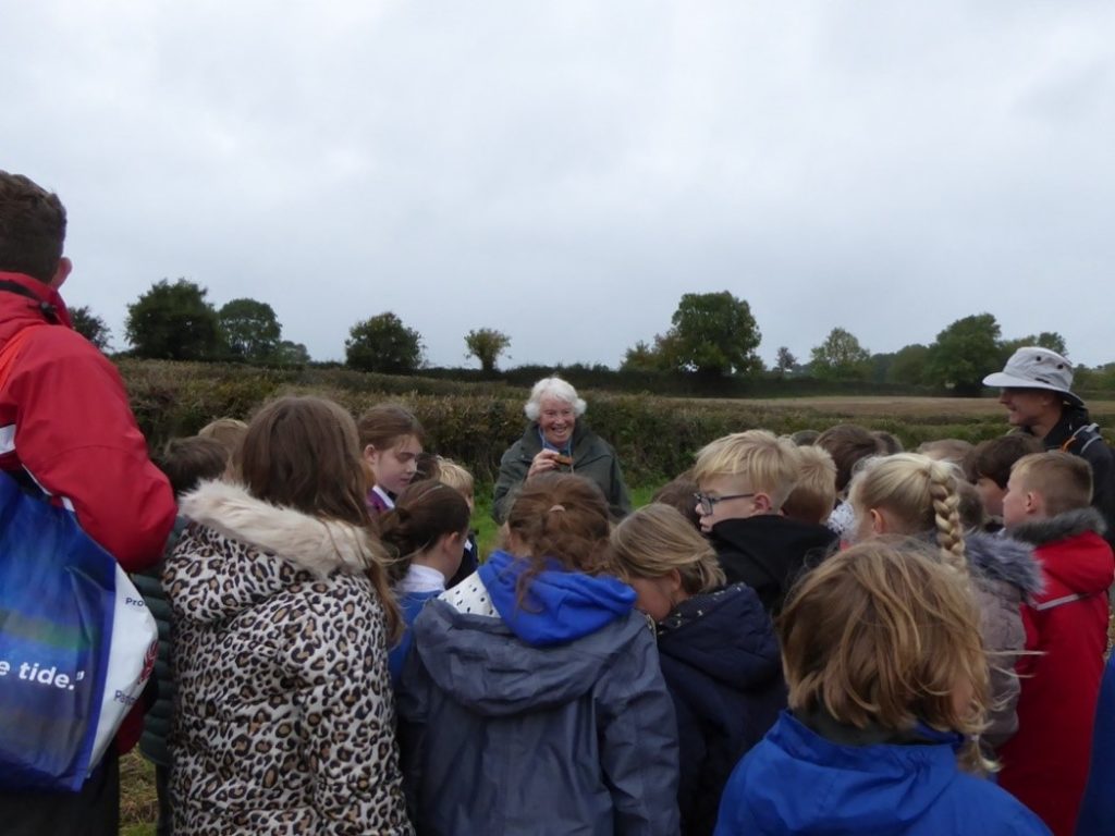 A group of school children listening to a lady talking, in a field