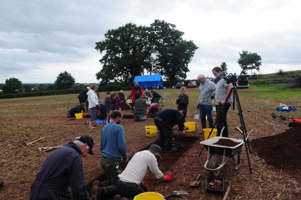 An archaeological excavation in a view with large trees in the background. People can be seen kneeling and trowelling on the ground, with others standing and looking on